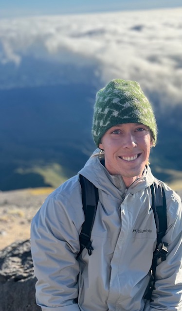 man in grey jacket and green patterned handknit hat with clouds in background, atop volcano